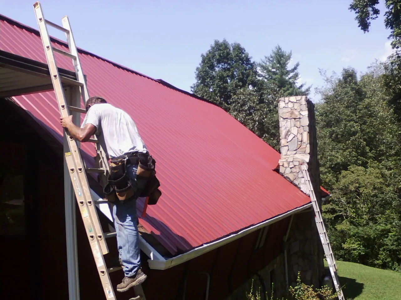 Roofer on a ladder beside a red metal roof and stone chimney — Soffit & Fascia Repair by Roofing Brothers in Mount Sterling
